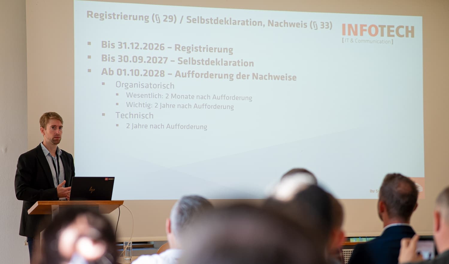 A presenter at the lectern beside a slide on NIS registration and self-declaration deadlines
