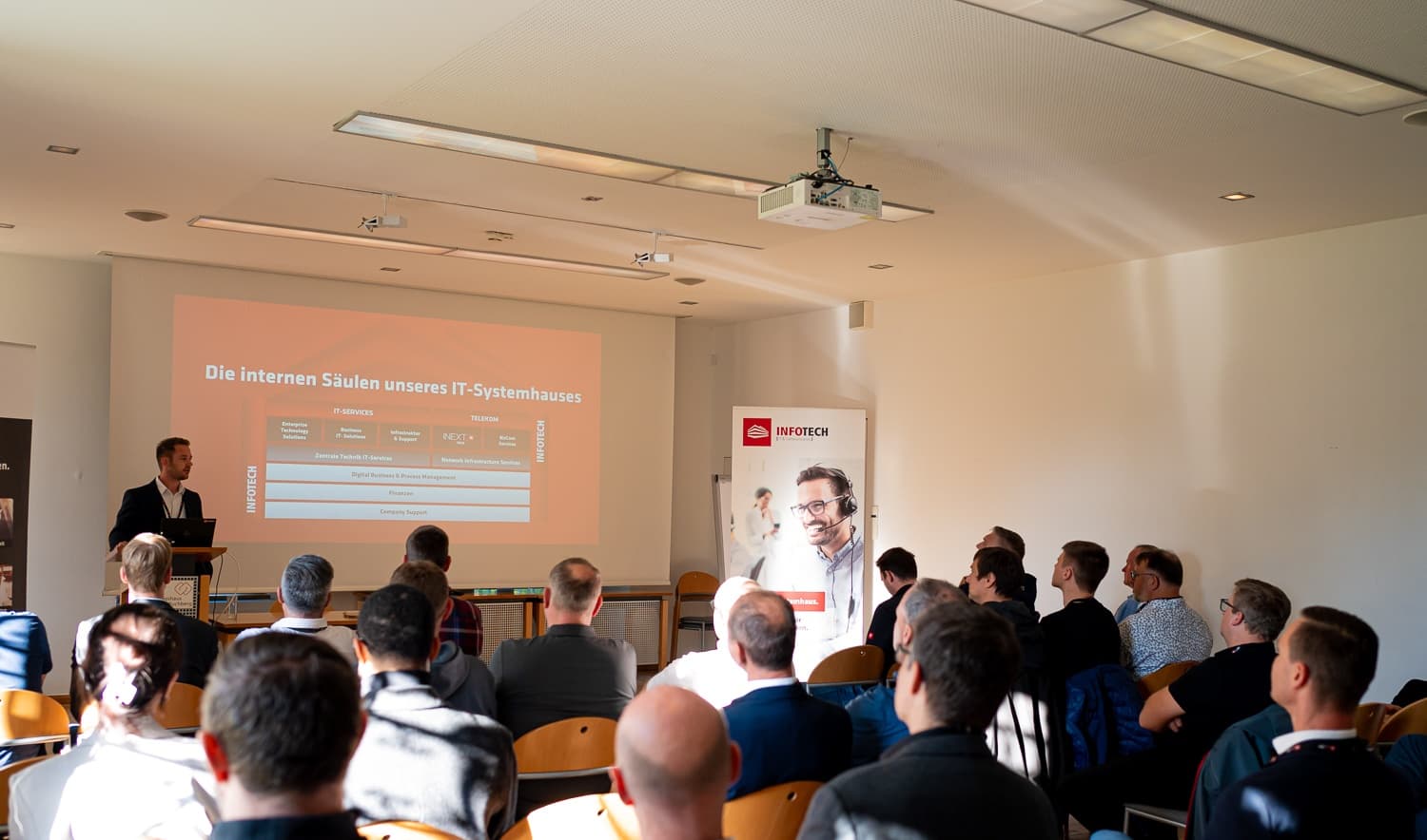 A presenter at the lectern beside a slide on the pillars of an IT systems house, seen from behind the audience in the Hofsaal of Schloss Puchberg