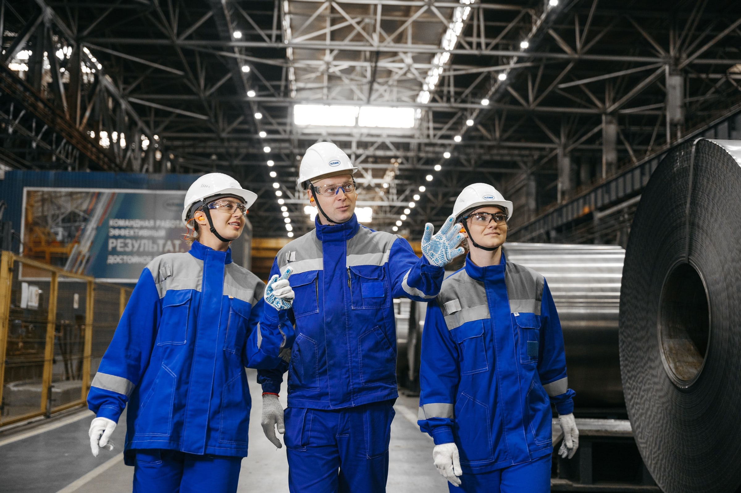 Three engineers in hard hats and blue overalls walking a steel coil plant floor, one pointing ahead